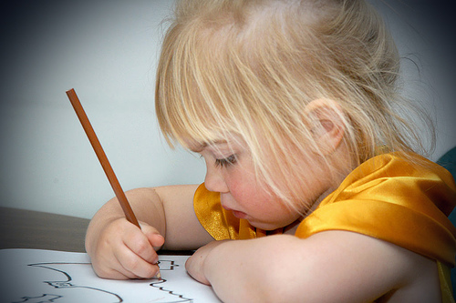 A blonde girl uses a brown Colorsed pencil to Colors in an image of a bat