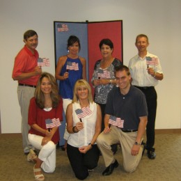 A group of men and women holding small American Flags pose in front of a red and blue diplay board