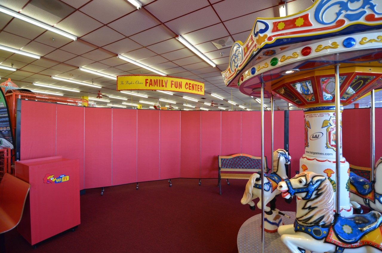 Two red Screenflex Room Dividers connected to create a curved barrier wall between a merry go round and a cafeteria at a family fun center.