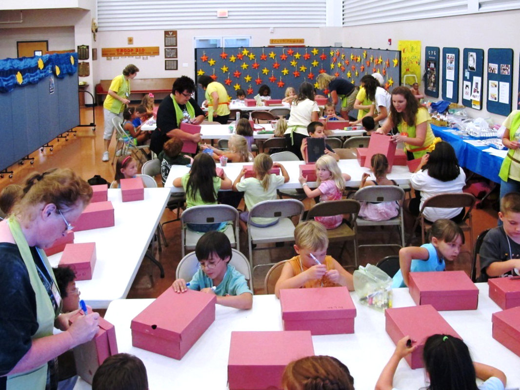 Adult staff assist students at a Vacation Bible School draw on red shoe boxes while sitting at long tables.