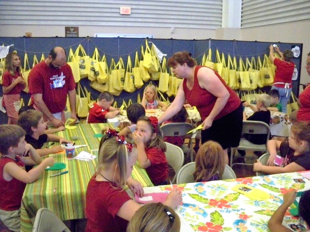 Yellow book bags hang on the sides of Screenflex Room Dividers while adults help students complete VBS crafts.
