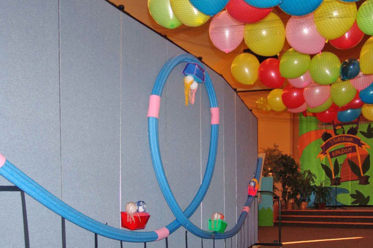 A roller coaster made from pool tubes is tacked to a Screenflex Room Dividers. Balloons rest on nets hanging from the ceiling.