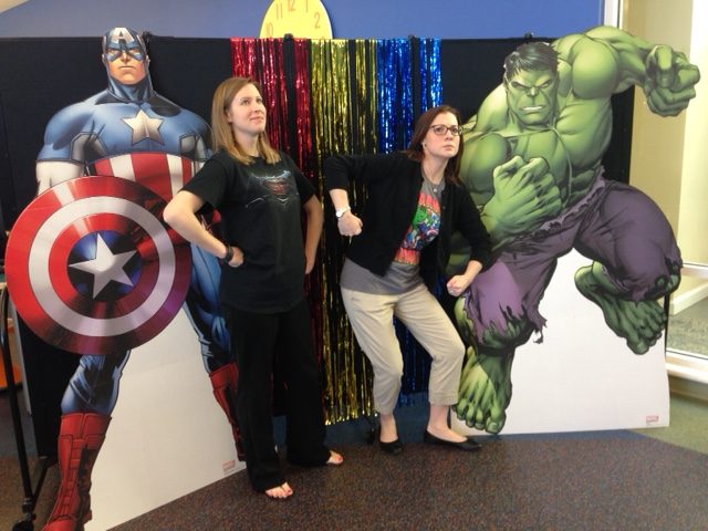 Life-size posters of superheroes flank the sides two librarians posing in front of a black room divider