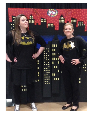 A pair of female librarians with batman t-shirts pose in front of a painted cartoon skyline