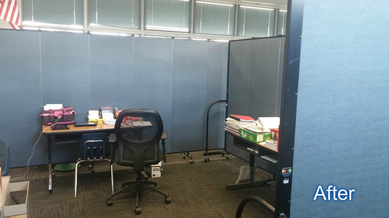 A set of student desks with supplies are arranged next to a portable wall that divides a classroom in two